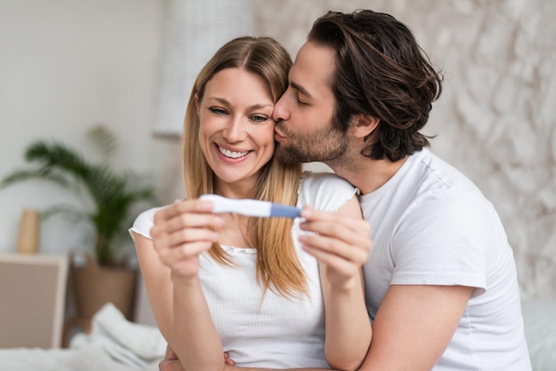 Casal jovem apaixonado se sentindo feliz com o teste de gravidez positivo beijando e abraçando em casa