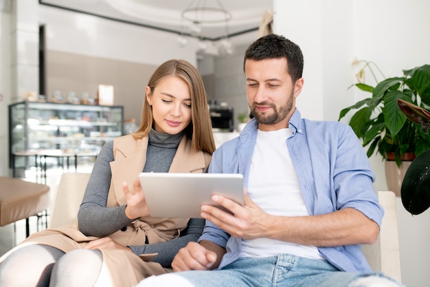 Casal jovem casual sentado em um sofá confortável e rolando no touchpad enquanto relaxa em um café aconchegante