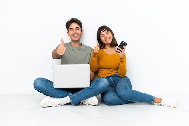 Casal jovem com um laptop e um celular sentado no chão fazendo um gesto de polegar para cima porque algo bom aconteceu