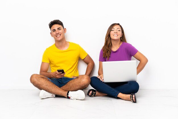 Casal jovem com um laptop sentado no chão, posando com os braços na cintura e sorrindo