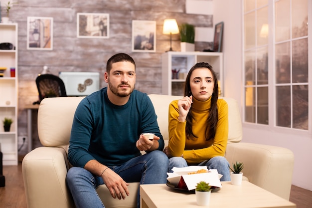 Casal jovem comendo frango frito em frente à tv na sala de estar