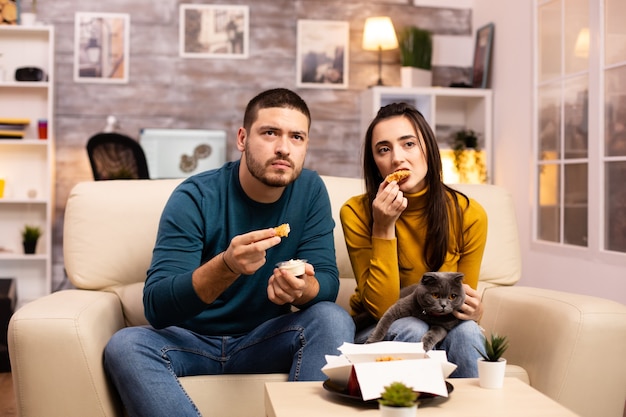Casal jovem comendo frango frito em frente à TV na sala de estar