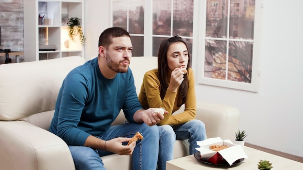 Casal jovem concentrado comendo frango frito enquanto assiste tv. casal comendo junk food.