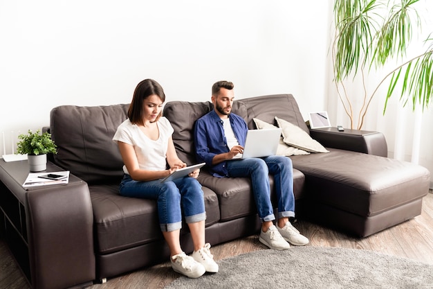 Casal jovem concentrado em roupas casuais, sentado em um sofá confortável e usando dispositivos enquanto trabalhava como freelancers em casa