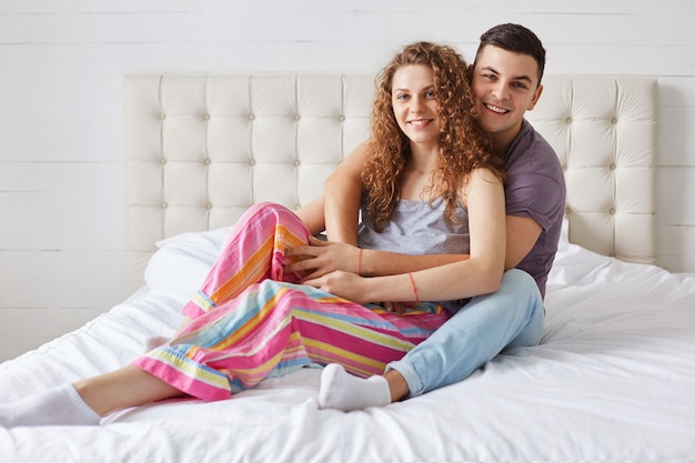 Casal jovem da família sentar e abraçar na cama confortável, desfrutar de juntos e bom dia, ter expressões positivas, usar pijama. mulher bonita encaracolada passa o tempo livre no quarto com o amante