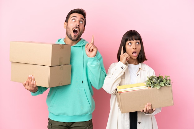 Casal jovem de beleza se mudando para uma nova casa entre caixas isoladas em fundo rosa, pensando em uma ideia apontando o dedo para cima