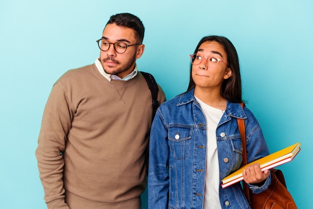 Casal jovem de estudante de raça mista isolado em fundo azul confuso, sente-se em dúvida e inseguro.