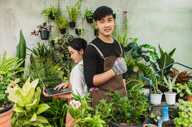 Casal jovem de jardineiros asiáticos usando avental usa equipamento de jardim e um laptop para pesquisar e cuidar das plantas da casa na estufa