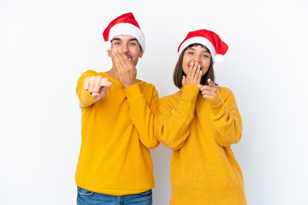 Casal jovem de raça mista celebrando o Natal isolado no fundo branco apontando com o dedo para alguém e rindo