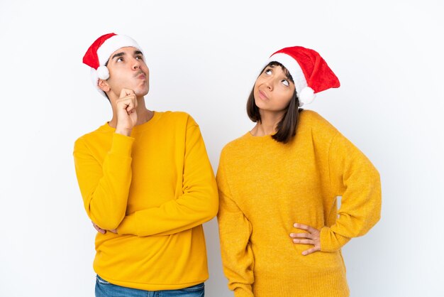 Casal jovem de raça mista celebrando o Natal isolado no fundo branco pensando uma ideia enquanto coça a cabeça