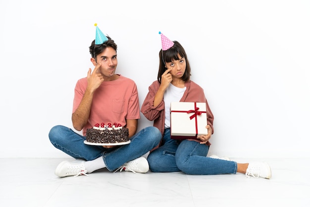 Casal jovem de raça mista comemorando aniversário, sentado no chão, isolado no fundo branco, olhando para a frente