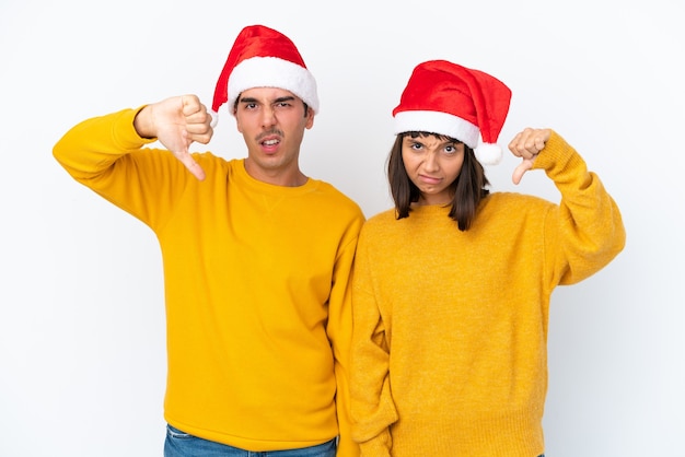 Casal jovem de raça mista comemorando o Natal isolado no fundo branco, mostrando o polegar para baixo com as duas mãos