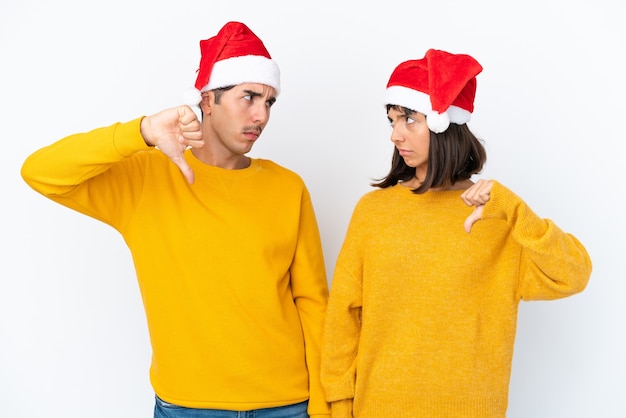 Casal jovem de raça mista comemorando o natal isolado no fundo branco, mostrando o sinal de polegar para baixo com expressão negativa