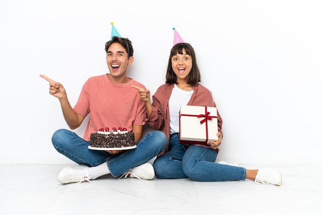 Casal jovem de raça mista comemorando um aniversário sentado no chão isolado no fundo branco apontando o dedo para o lado e apresentando um produto