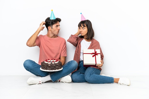 Casal jovem de raça mista comemorando um aniversário sentado no chão isolado no fundo branco com dúvidas enquanto coça a cabeça