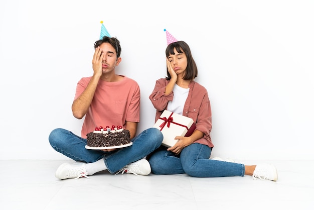 Casal jovem de raça mista comemorando um aniversário sentado no chão isolado no fundo branco com surpresa e expressão facial chocada