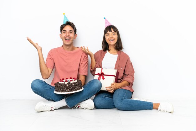 Casal jovem de raça mista comemorando um aniversário sentado no chão isolado no fundo branco com surpresa e expressão facial chocada