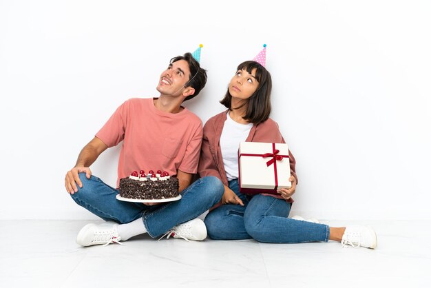 Casal jovem de raça mista comemorando um aniversário sentado no chão isolado no fundo branco olhando para cima enquanto sorria