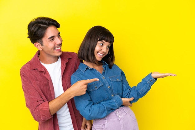 Casal jovem de raça mista isolado em fundo amarelo apontando para trás e apresentando um produto