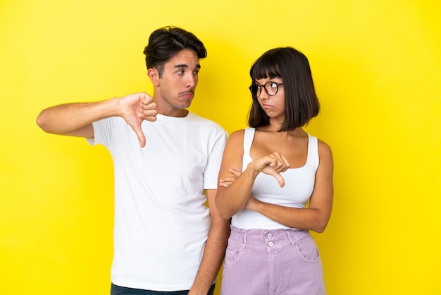 Casal jovem de raça mista isolado em fundo amarelo, mostrando o polegar para baixo com expressão negativa