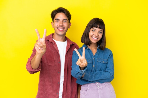 Casal jovem de raça mista isolado em fundo amarelo sorrindo e mostrando sinal de vitória