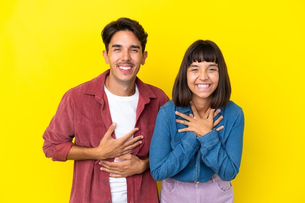 Casal jovem de raça mista isolado em fundo amarelo sorrindo muito enquanto coloca as mãos no peito