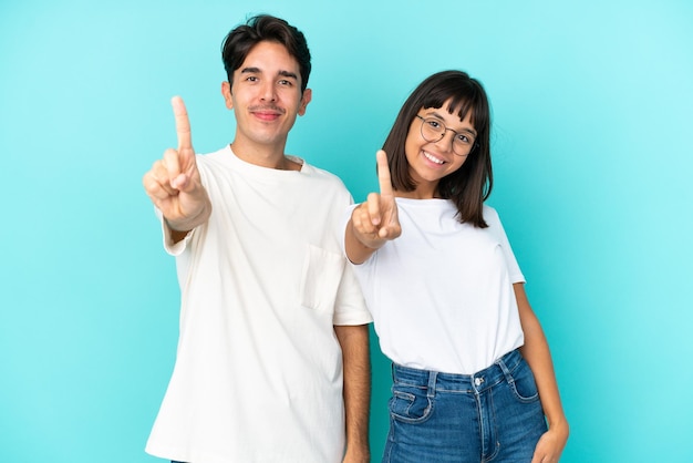 Casal jovem de raça mista isolado em fundo azul mostrando e levantando um dedo