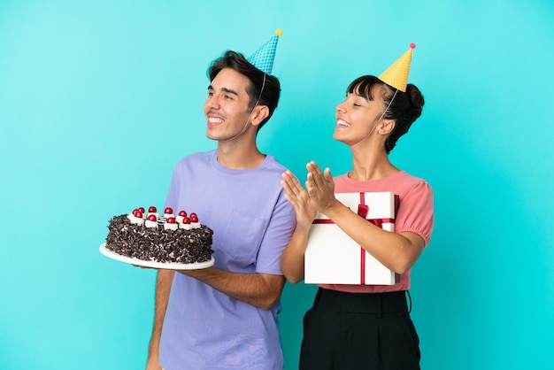 Casal jovem de raça mista segurando bolo de aniversário e presente isolado em fundo azul aplaudindo após apresentação em uma conferência