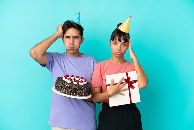 Casal jovem de raça mista segurando bolo de aniversário e presente isolado em fundo azul com uma expressão de frustração e não compreensão