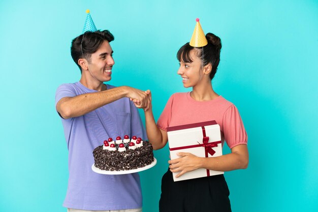 Casal jovem de raça mista segurando bolo de aniversário e presente isolado no aperto de mão de fundo azul depois de um bom negócio