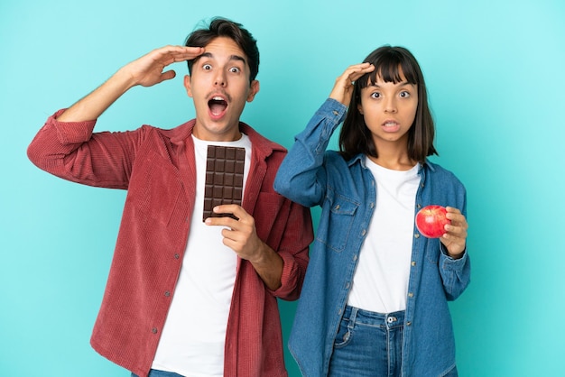 Casal jovem de raça mista segurando maçã e chocolate isolado em fundo azul acaba de perceber algo e pretende a solução