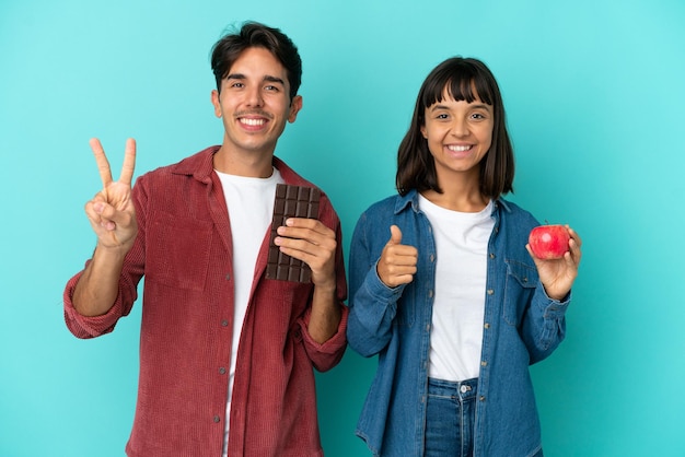 Casal jovem de raça mista segurando maçã e chocolate isolado no fundo azul, dando um polegar para cima gesto com as duas mãos e sorrindo