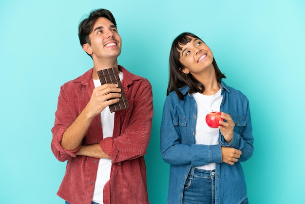 Casal jovem de raça mista segurando maçã e chocolate isolados no fundo azul, olhando para cima enquanto sorri