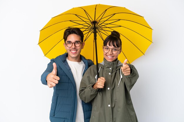 Casal jovem de raça mista segurando um guarda-chuva isolado no fundo branco dando um polegar para cima gesto porque algo bom aconteceu