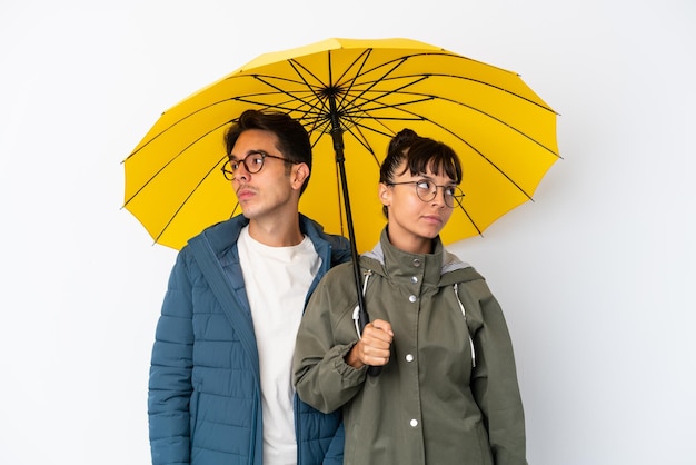 Casal jovem de raça mista segurando um guarda-chuva isolado no fundo branco nervoso e assustado