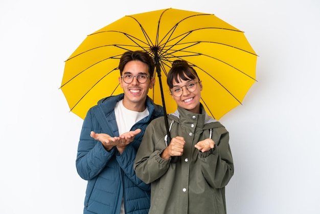 Casal jovem de raça mista segurando um guarda-chuva isolado no fundo branco segurando copyspace imaginário na palma da mão para inserir um anúncio