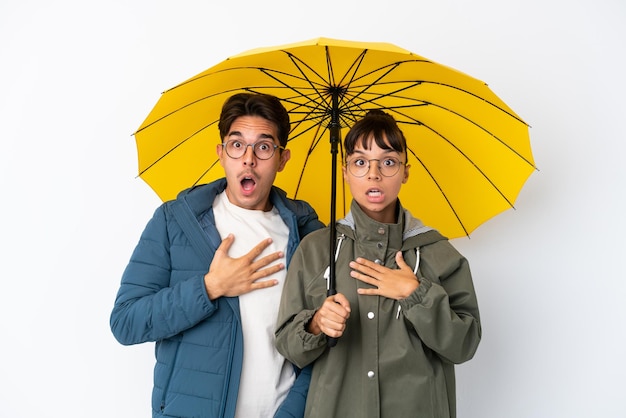 Casal jovem de raça mista segurando um guarda-chuva isolado no fundo branco surpreso e chocado ao olhar para a direita
