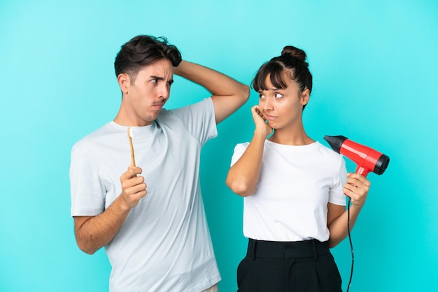 Casal jovem de raça mista segurando um secador de cabelo e escova de dentes isolado em fundo azul com dúvidas enquanto coça a cabeça