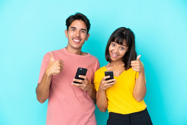 Casal jovem de raça mista segurando um telefone celular isolado em um fundo azul, fazendo um gesto de polegar para cima com as duas mãos e sorrindo