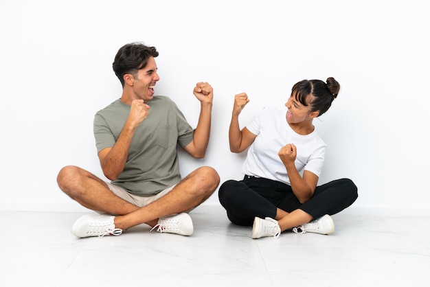 Casal jovem de raça mista sentado no chão isolado no fundo branco comemorando uma vitória na posição de vencedor