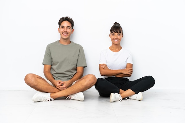 Casal jovem de raça mista sentado no chão isolado no fundo branco mantendo os braços cruzados em posição frontal