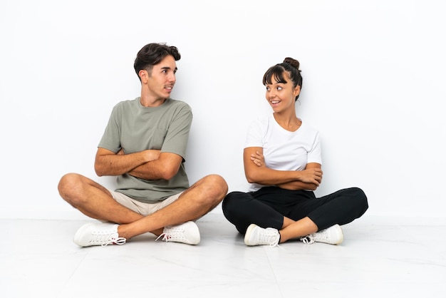 Casal jovem de raça mista sentado no chão isolado no fundo branco olhando por cima do ombro com um sorriso