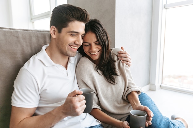 Casal jovem e adorável segurando copos enquanto está sentado no chão no sofá em casa