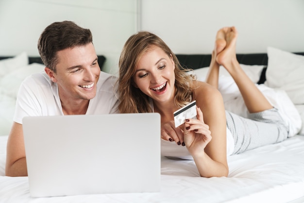 Casal jovem e atraente e feliz deitado na cama no quarto em casa, usando o laptop para fazer compras