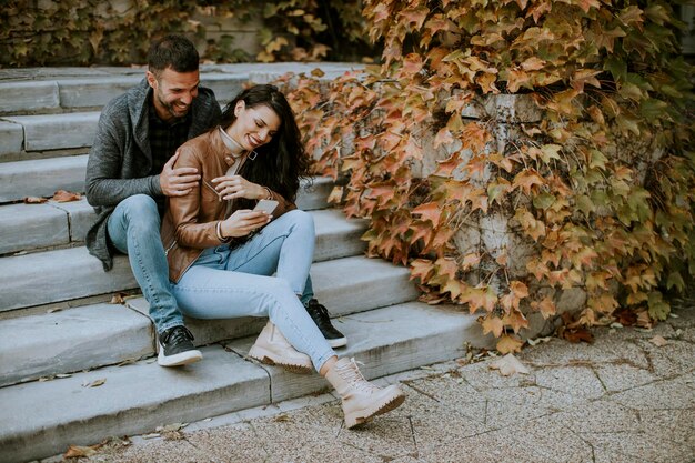 Casal jovem e bonito sentado em uma escada externa em um dia de outono e usando um telefone celular