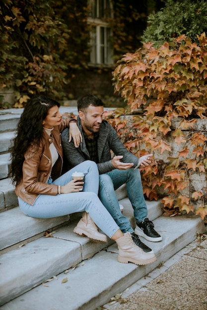 Casal jovem e bonito sentado em uma escada externa em um dia de outono e usando um telefone celular