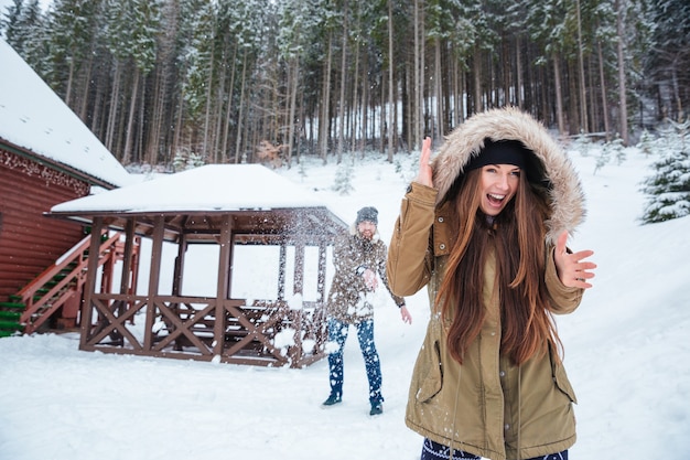 Casal jovem e feliz e animado jogando bolas de neve e gritando perto de casa na floresta de inverno