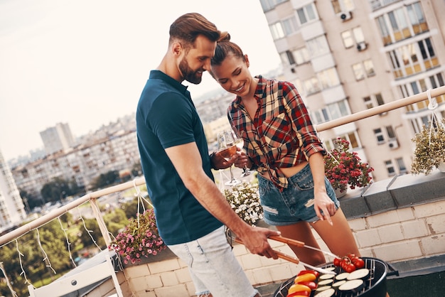 Casal jovem e fofo em roupas casuais, preparando um churrasco e sorrindo em pé no pátio da cobertura ao ar livre