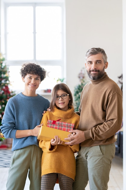 Casal jovem e sua linda filha adolescente segurando caixas de presentes com presentes enquanto estão de pé contra uma árvore de natal decorada na sala de estar