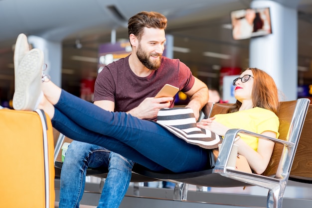 Casal jovem em camisetas coloridas sentado com bagagens na sala de espera do aeroporto